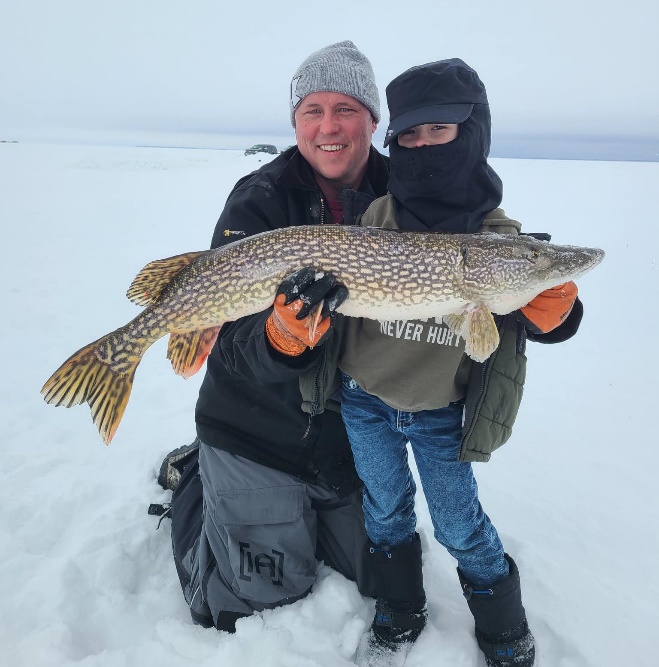 Joel Peterson ice fishing on a frozen Minnesota lake, holding up a large northern pike alongside a young angler